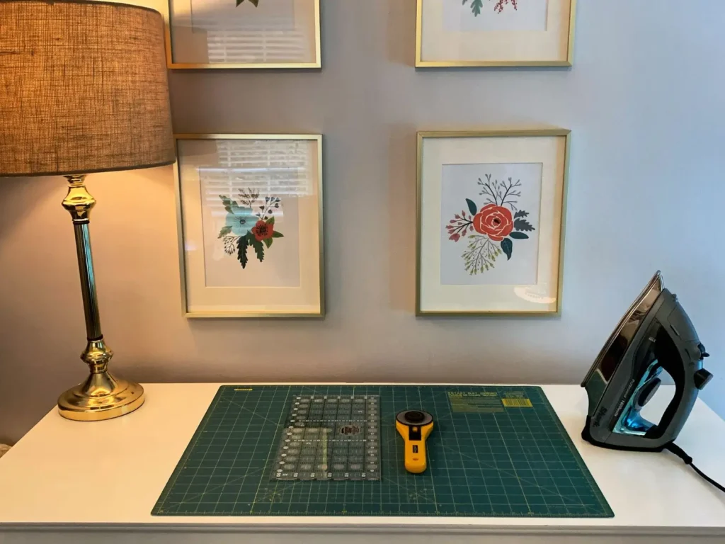 Quilting cutting station with quilting tools: a green cutting mat, rotary cutter, and ruler on a white dresser, surrounded by framed floral artwork, a table lamp, and an iron