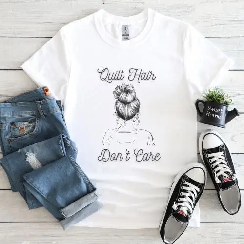 White t-shirt with a sketch of a woman’s messy bun hairstyle and the text 'Quilt Hair Don’t Care,' styled with folded jeans, black Converse sneakers, and farmhouse decor on a light wooden surface.