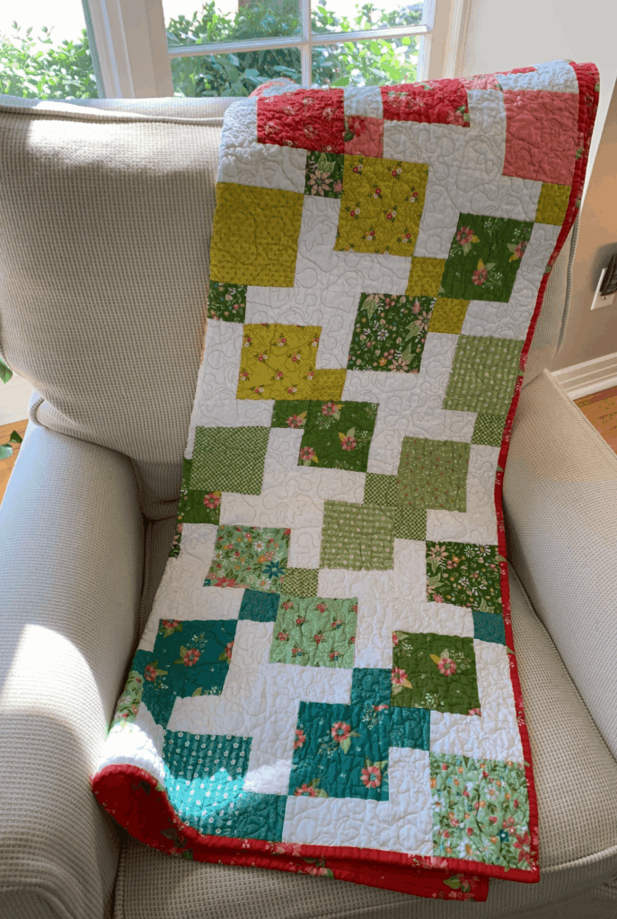 Color wash disappearing nine-patch baby quilt draped over a light upholstered chair near a window, showing green, teal, yellow, and coral blocks with red binding.