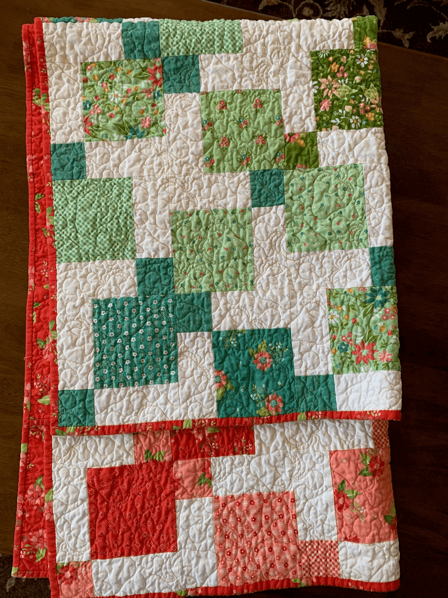 A folded Color Wash Disappearing 9-Patch quilt featuring green, teal, and coral floral fabrics with red binding, displayed on a wooden surface.