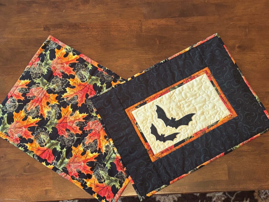 Two Halloween quilted placemats displayed on a wooden table — one with black bats on cream and orange borders, the other with bright fall leaf print backing.