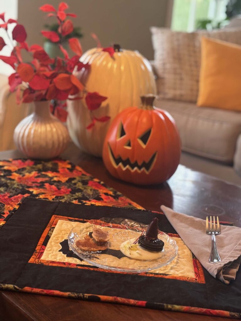Quilted Halloween placemat with bat appliqué styled on a wooden table with pumpkins, orange leaves, and a small plate of Halloween treats.