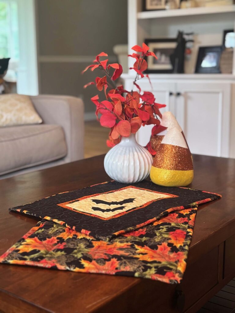 Two quilted Halloween placemats stacked on a wooden table, surrounded by autumn décor including a white vase with red leaves and a glittery candy-corn ornament.