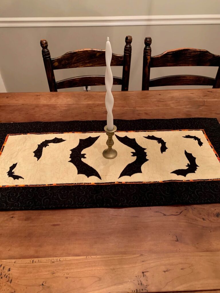 Quilted Halloween table runner featuring black bat appliqués on a cream background with orange and black borders, displayed on a wooden dining table with a twisted white taper candle in a brass holder.