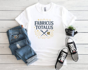 White t-shirt with the phrase “Fabricus Totalus” in a magical typography design, featuring crossed scissors and a quilt block, styled with jeans and black sneakers on a light wood background.