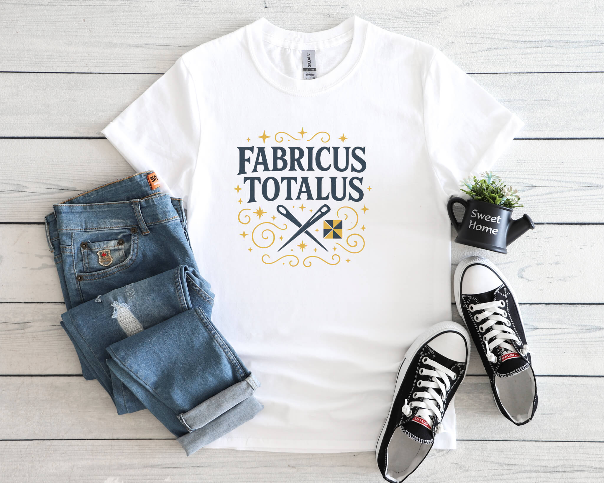 White t-shirt with the phrase “Fabricus Totalus” in a magical typography design, featuring crossed scissors and a quilt block, styled with jeans and black sneakers on a light wood background.