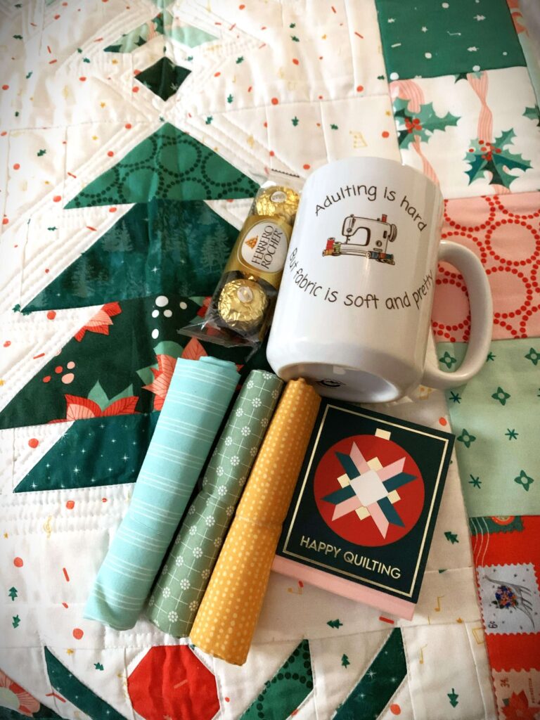 Mini quilt swap gifts including a mug, rolled fabric, chocolates, and a notepad arranged on a quilt.
