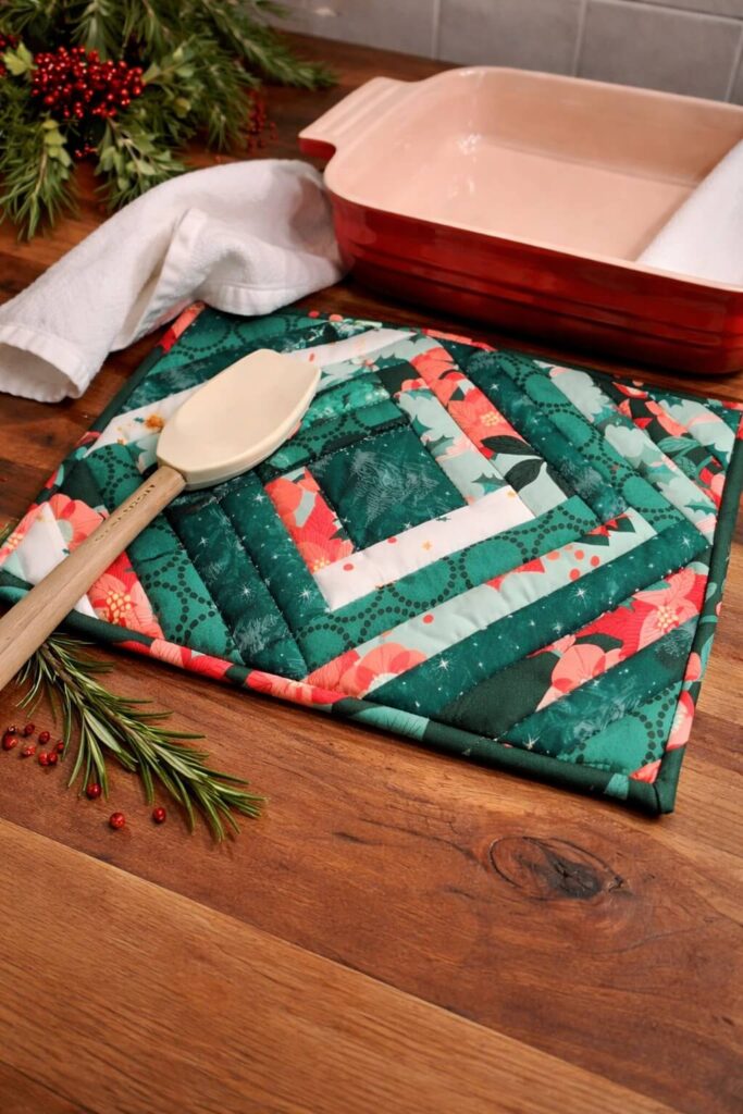 Quilted casserole hot pad in green, teal, and red fabrics on a wooden countertop with baking dish and spatula