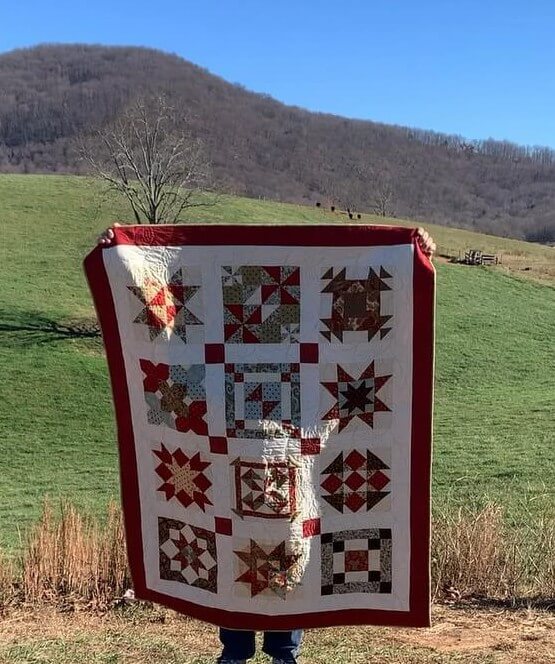 Finished Christmas Block of the Month quilt displayed outdoors in the mountains, featuring red and cream sashing, star blocks, and a deep red border.