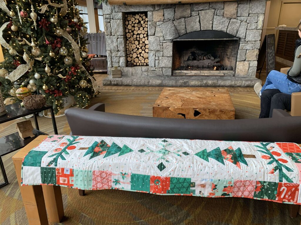 Christmas quilted table runner displayed on a bench in front of a stone fireplace and decorated Christmas tree, featuring green, white, and red patchwork fabrics.