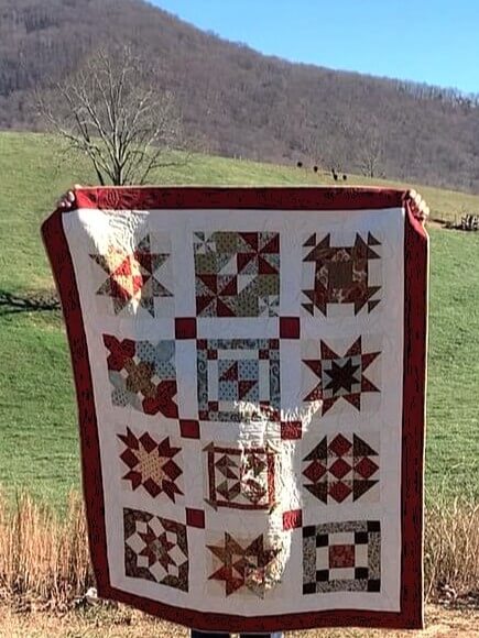 Handmade sampler quilt with red binding and classic block designs, photographed outdoors against rolling green hills and a blue sky.
