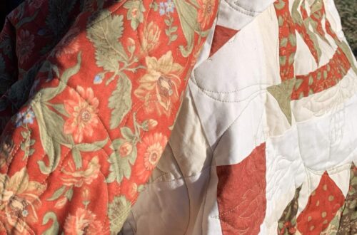 Close-up of a handmade sampler quilt with warm red and cream patchwork, shown wrapped around a child outdoors in natural light.
