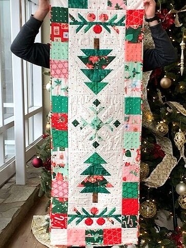 Under the Pines Christmas table runner held vertically in front of a decorated Christmas tree.